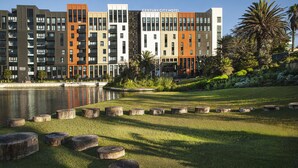 Ein modernes Hotel mit mehreren Etagen, einem angelegten Garten mit Holz-Pflanzgefäßen und einem Gewässer im Vordergrund.