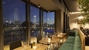 Ein Restaurant mit Blick auf die nächtliche Skyline, das große Fenster, Holztische und grüne Sitzmöbel bietet.
