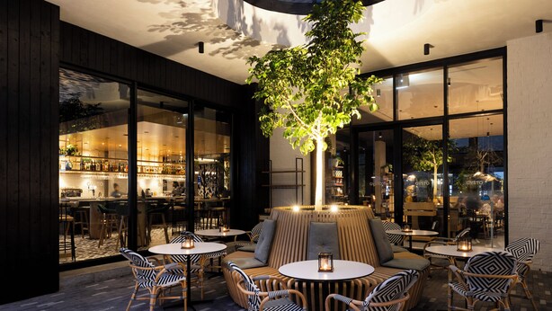 Ein Außenbereich mit runden Tischen und Korbstühlen, umgeben von großen Fenstern und einem zentralen Baum.