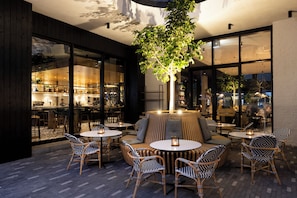 An outdoor seating area with round tables and wicker chairs, surrounded by large windows and a central tree.