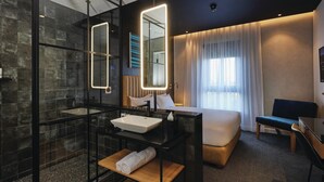 Ein modernes Hotelzimmer mit Duschkabine aus Glas, einem großen Spiegel, einem Nachttisch mit Handtuch, einer Couch und einem Fernseher.