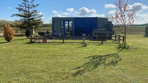 A blue modular building with a patio area, surrounded by a wooden fence, in a grassy field with trees and a clear sky.