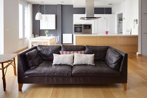 A modern living room with a dark leather sofa, two cushions, a wooden dining table, and a kitchen in the background.