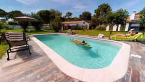 A swimming pool with a wooden deck, lounge chairs, and a table with a white cover. - 2 gites in a green setting (Sainte-Marie-de-Ré)