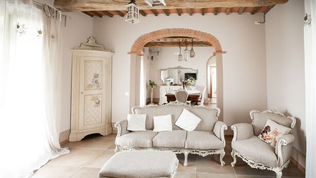 Um quarto com um sofá bege, uma poltrona combinando e um banquinho, um armário branco e um arco de tijolos.
