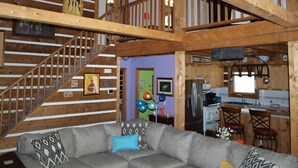 A spacious living area with a gray sectional sofa, wooden beams, a staircase, and a kitchen area with a refrigerator and stove. - Come to "LOG OUT" and have fun! (Pinnacle)