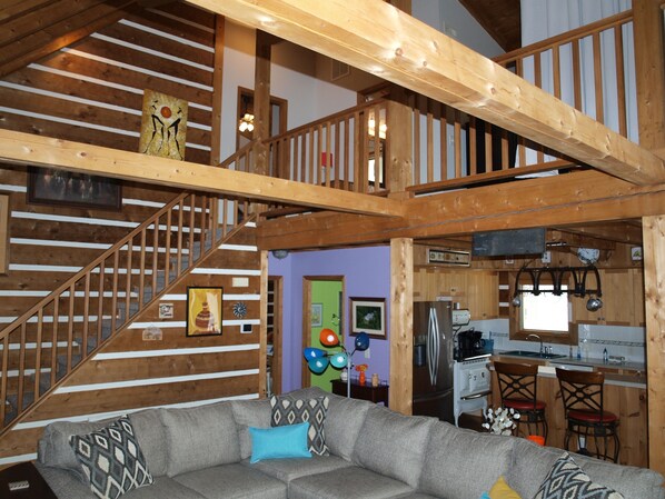 A spacious living area with a gray sectional sofa, wooden beams, a staircase, and a kitchen area with a refrigerator and stove.