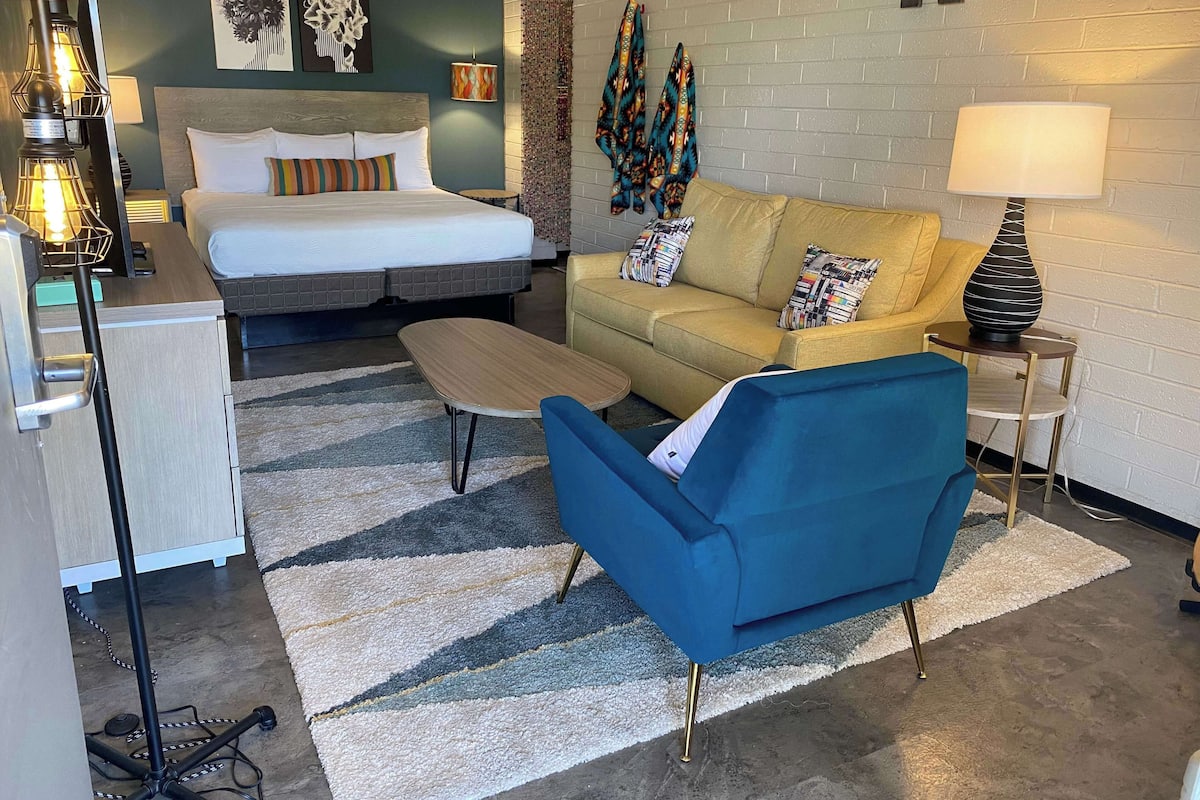 Habitación de hotel con cama, sofá, sillón azul, una mesita y una lámpara.