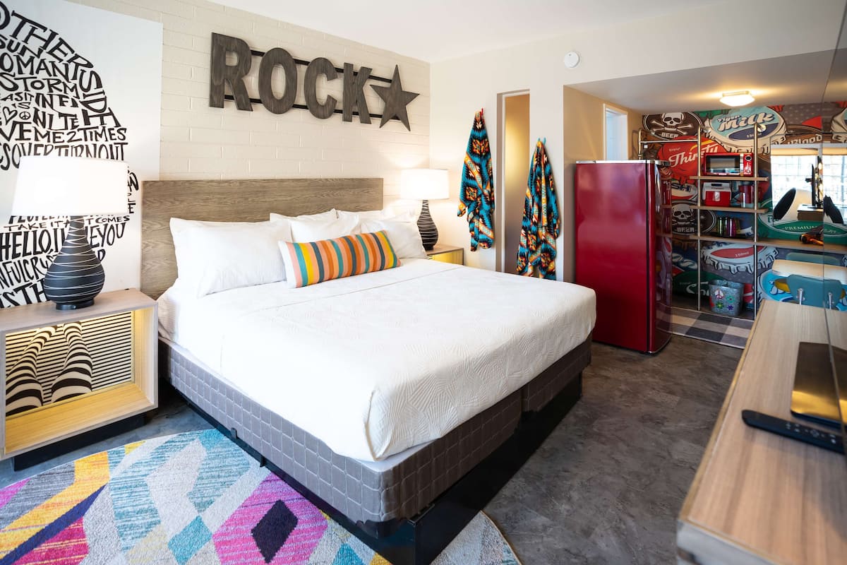 Una habitación de hotel moderna con una cama grande, un refrigerador rojo, una mesita de noche de madera y una pared con un letrero que dice 'ROCK'.