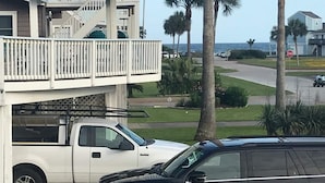 Un edificio de dos pisos con un balcón, una camioneta blanca estacionada adelante y palmeras al fondo.
