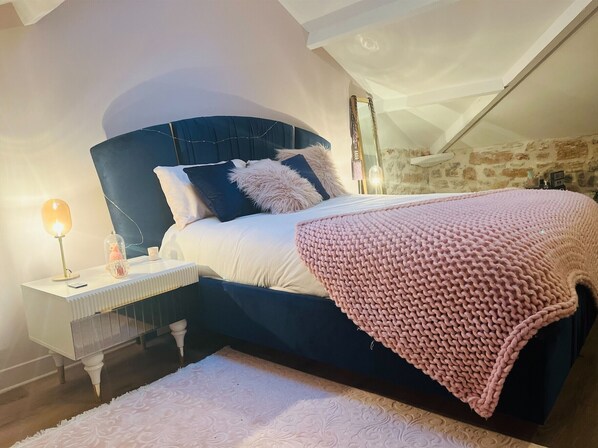 A bedroom with a dark blue headboard, white bedding, a pink knitted throw, and a bedside table with a lamp and decorative items.