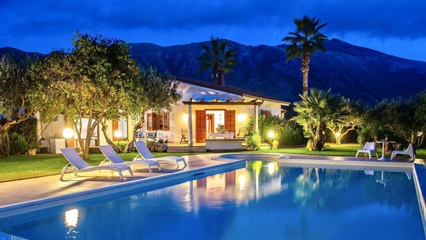 Una piscina con sillones de descanso, una casa con patio y montañas al fondo.