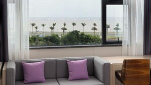 Camera d'hotel con un divano grigio, due cuscini viola, una sedia di legno e vista sulla spiaggia e sulle palme attraverso una finestra.
