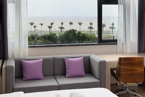 A hotel room with a grey sofa, two purple pillows, a wooden chair, and a view of the beach and palm trees through a window. - Lavande Hotel (Zhuhai Qinglv North Road Tangjiawan Beach) (Zhuhai)