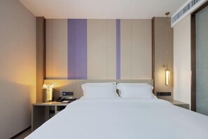 Quarto de hotel com uma cama grande, duas mesas de cabeceira, um criado-mudo, uma lâmpada e um ponto de luz fixado na parede.