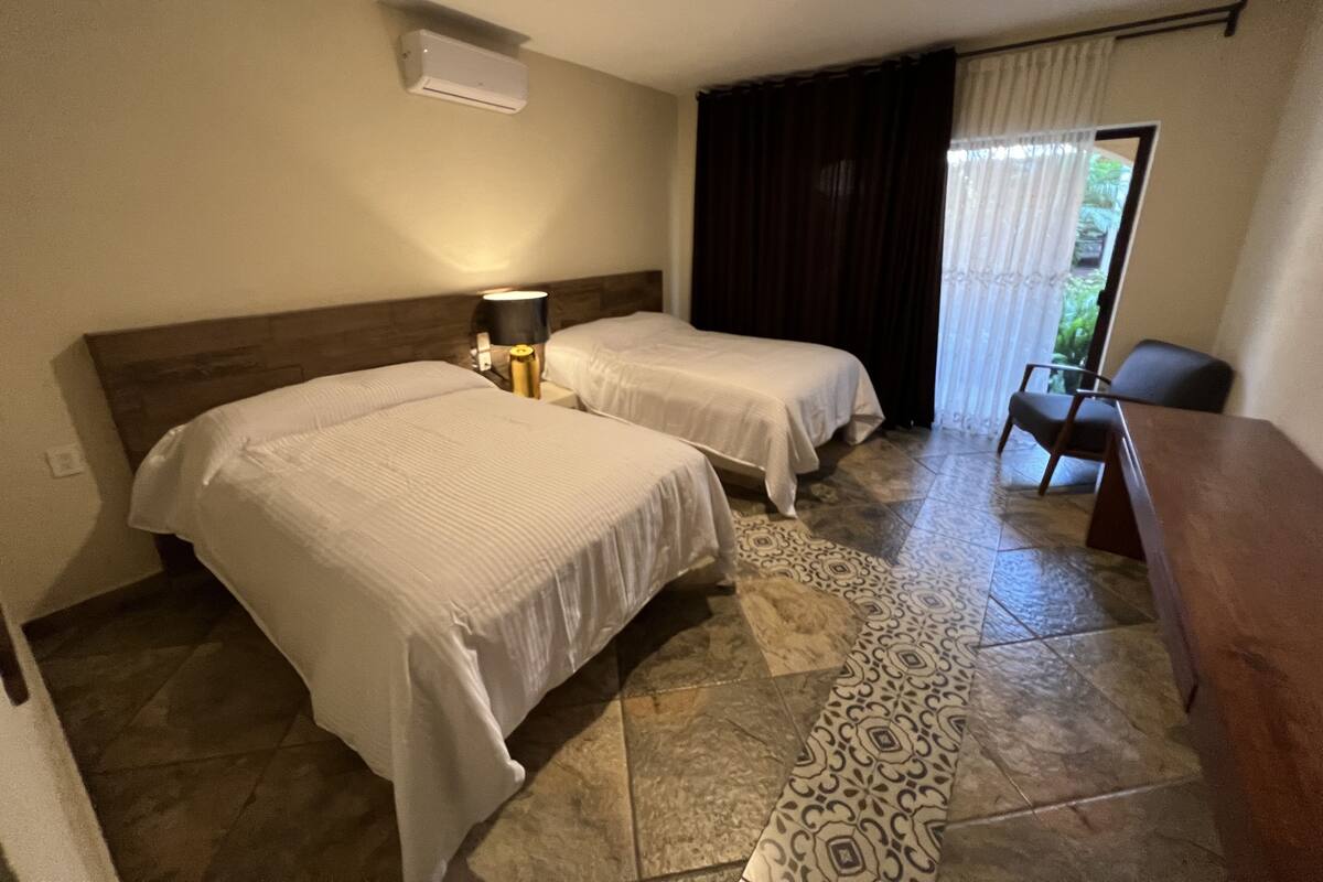A hotel room with two beds, a desk, a chair, a lamp, and a window with curtains.