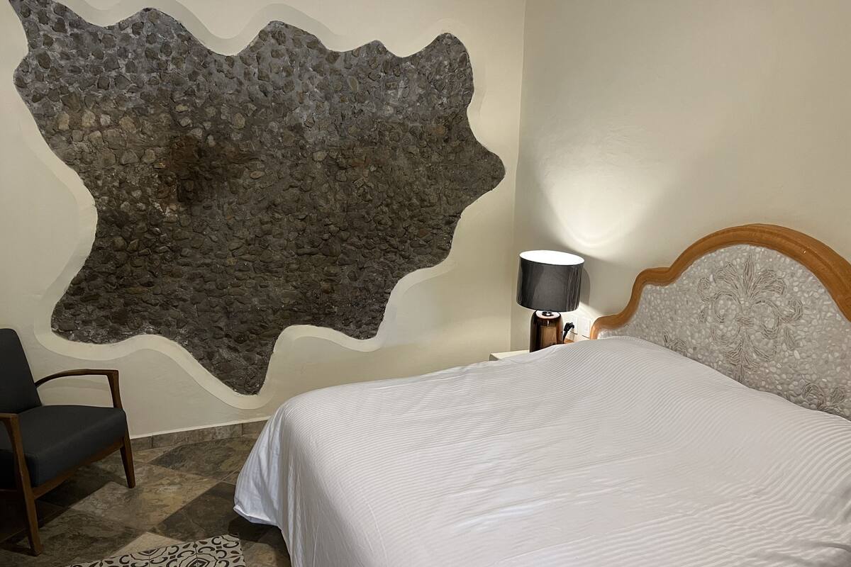 A bedroom with a stone wall, a bed, a chair, and a nightstand with a lamp.