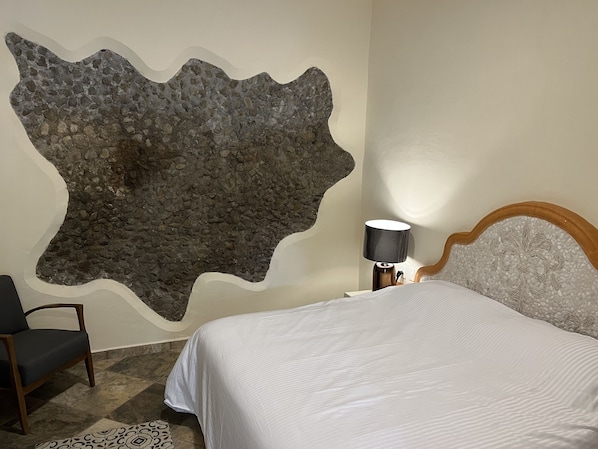 A bedroom with a stone wall, a bed, a chair, and a nightstand with a lamp. - Mesón de las Delicias (Cuernavaca)