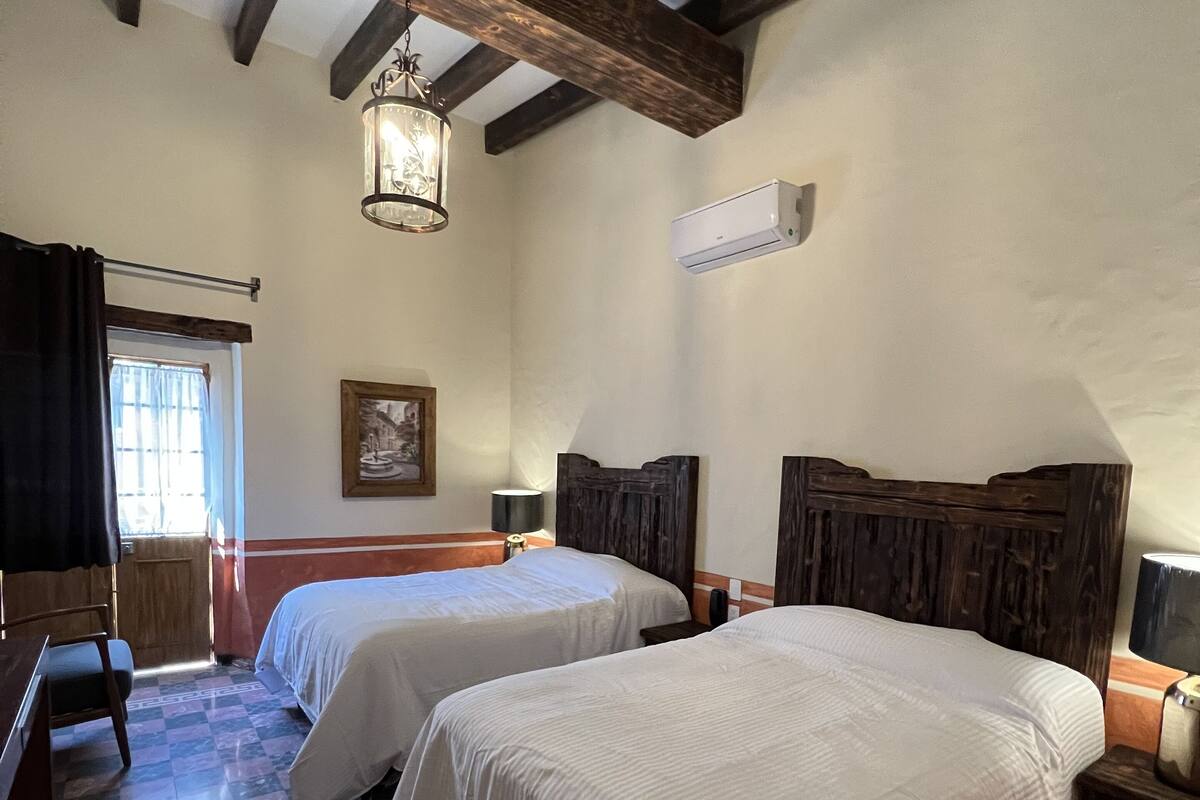 A room with two beds, a wooden ceiling, an air conditioning unit, a checkered floor, and a window with curtains.