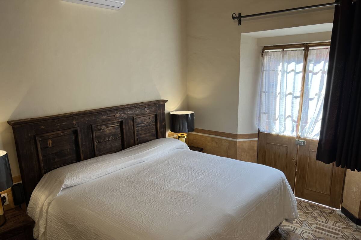 A bedroom with a large bed, a nightstand with a lamp, a wooden headboard, a window with curtains, and a patterned floor.