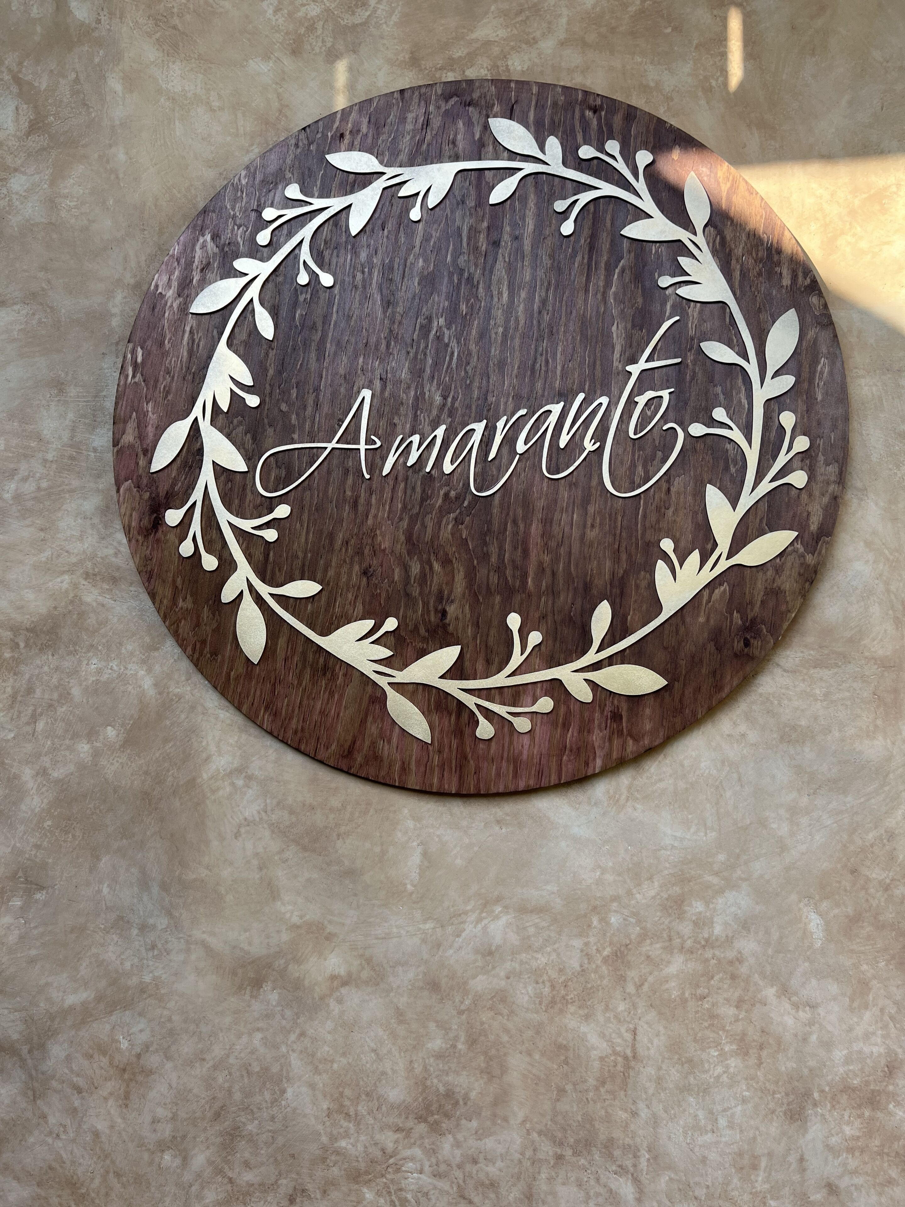 A circular wooden plaque with the name "Amaranto" surrounded by a decorative leaf pattern.