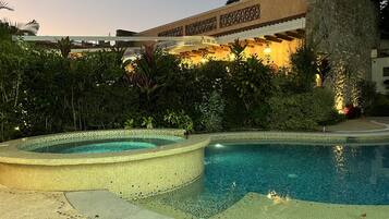 Una piscina con borde de azulejos, rodeada de exuberante vegetación y un edificio con paredes de piedra e iluminación decorativa.