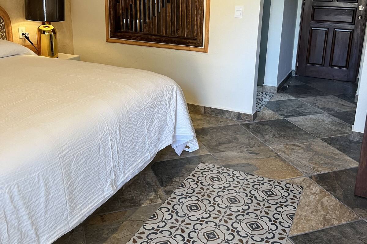 A bedroom with a bed, a patterned rug, a wall-mounted wooden panel, a nightstand with a lamp, and a door leading to another room.