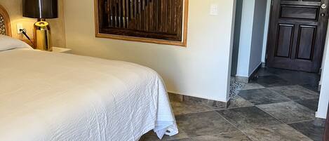 A bedroom with a bed, a patterned rug, a wall-mounted wooden panel, a nightstand with a lamp, and a door leading to another room.