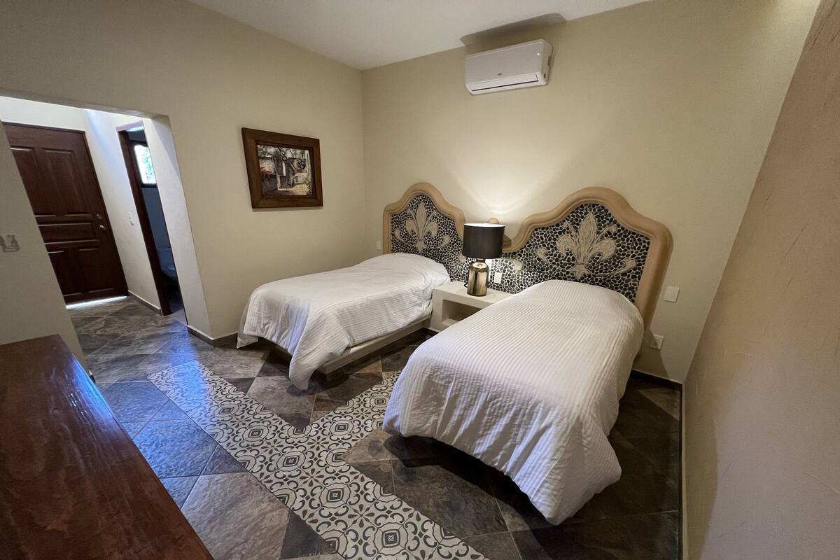 A hotel room with two beds, a nightstand with a lamp, a wall-mounted air conditioner, and a patterned rug.