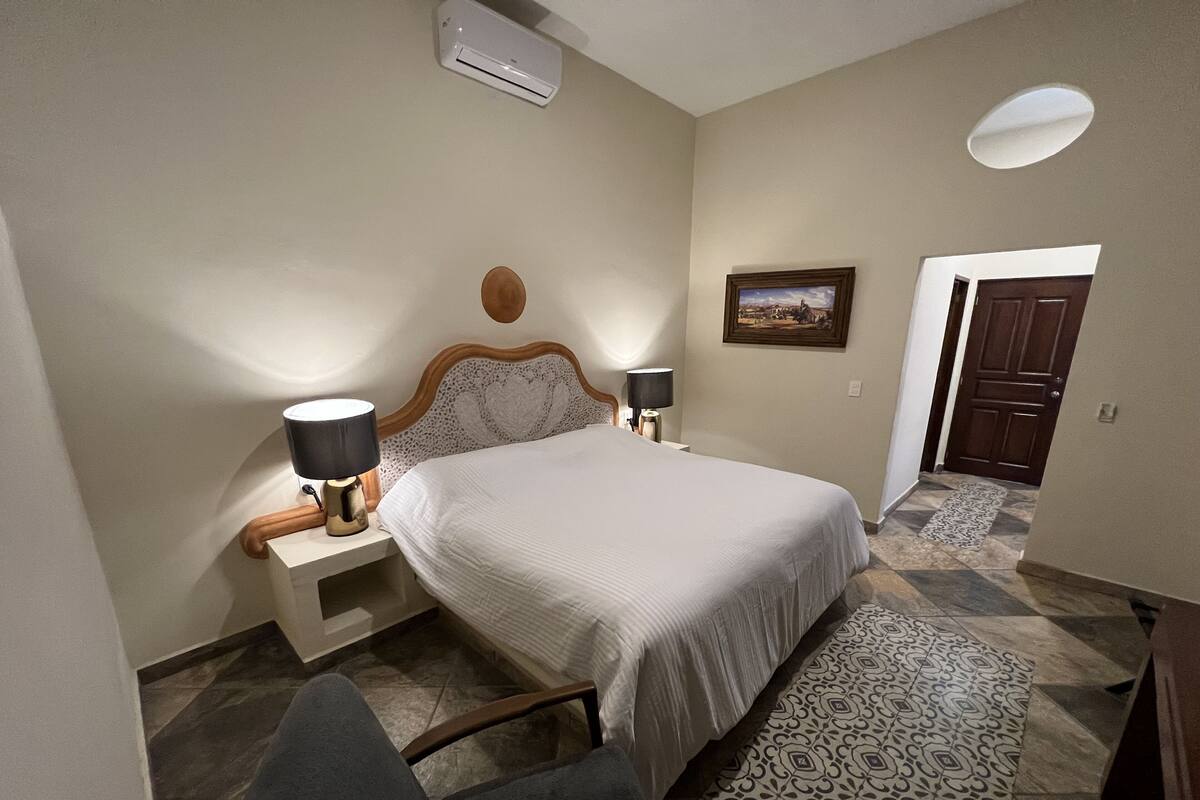 A bedroom with a bed, bedside lamps, a nightstand, a wall-mounted air conditioner, a door, and a patterned rug.