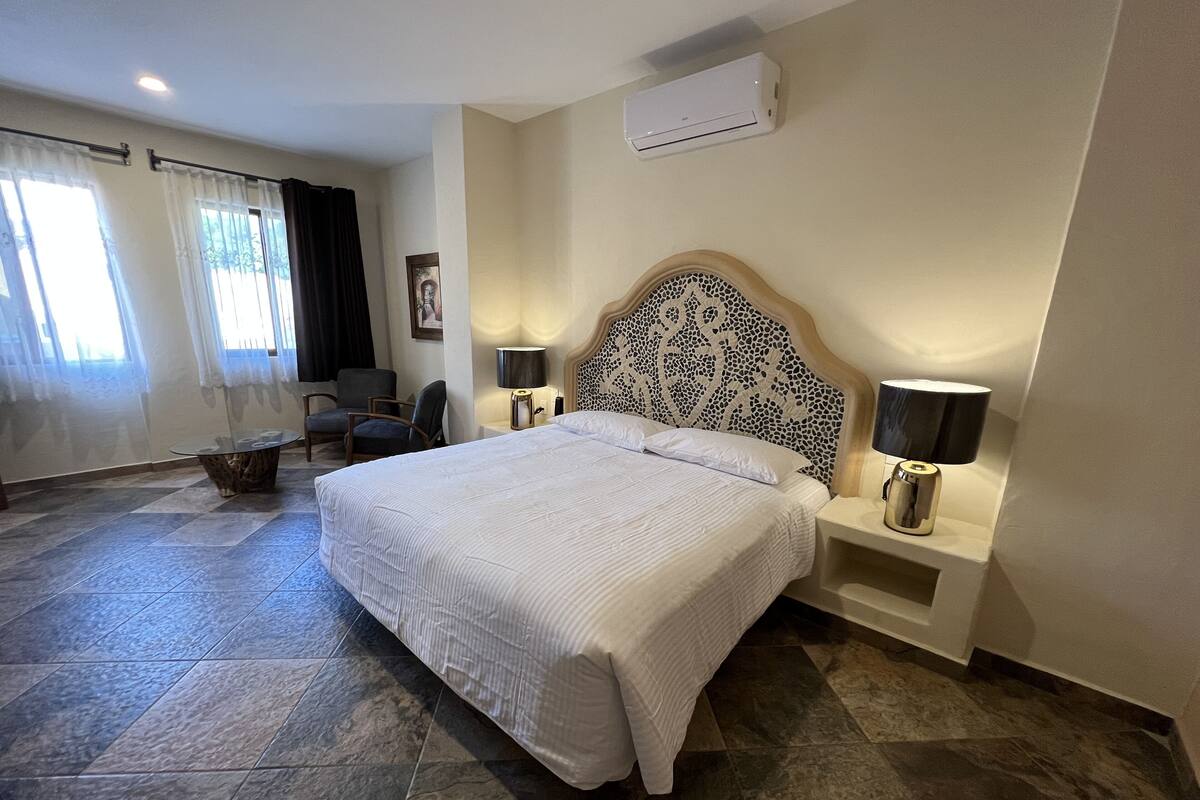 A hotel room with a large bed, two bedside tables with lamps, a chair, a small table, and a window with curtains.