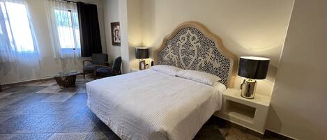 A hotel room with a large bed, two bedside tables with lamps, a chair, a small table, and a window with curtains.