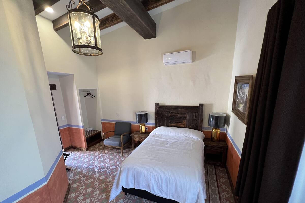 A hotel room with a large bed, a chair, a desk, a lamp, and a hanging light fixture.