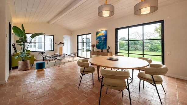 Une pièce spacieuse avec une table de salle à manger ronde, plusieurs chaises, une grande fenêtre donnant sur un paysage verdoyant et un luminaire suspendu.