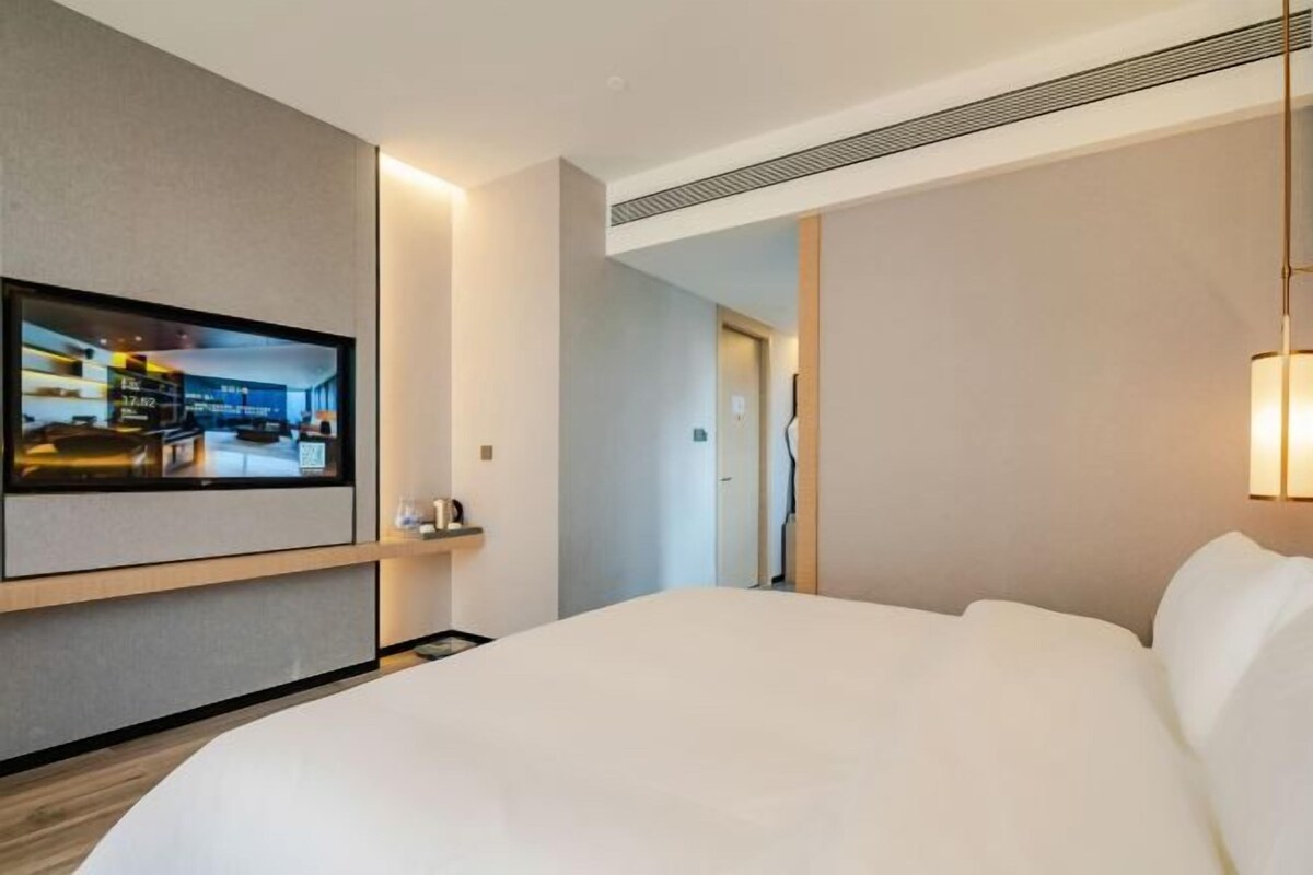 A hotel room with a large bed, a mounted TV, a bedside table with a lamp, and a door leading to another room.
