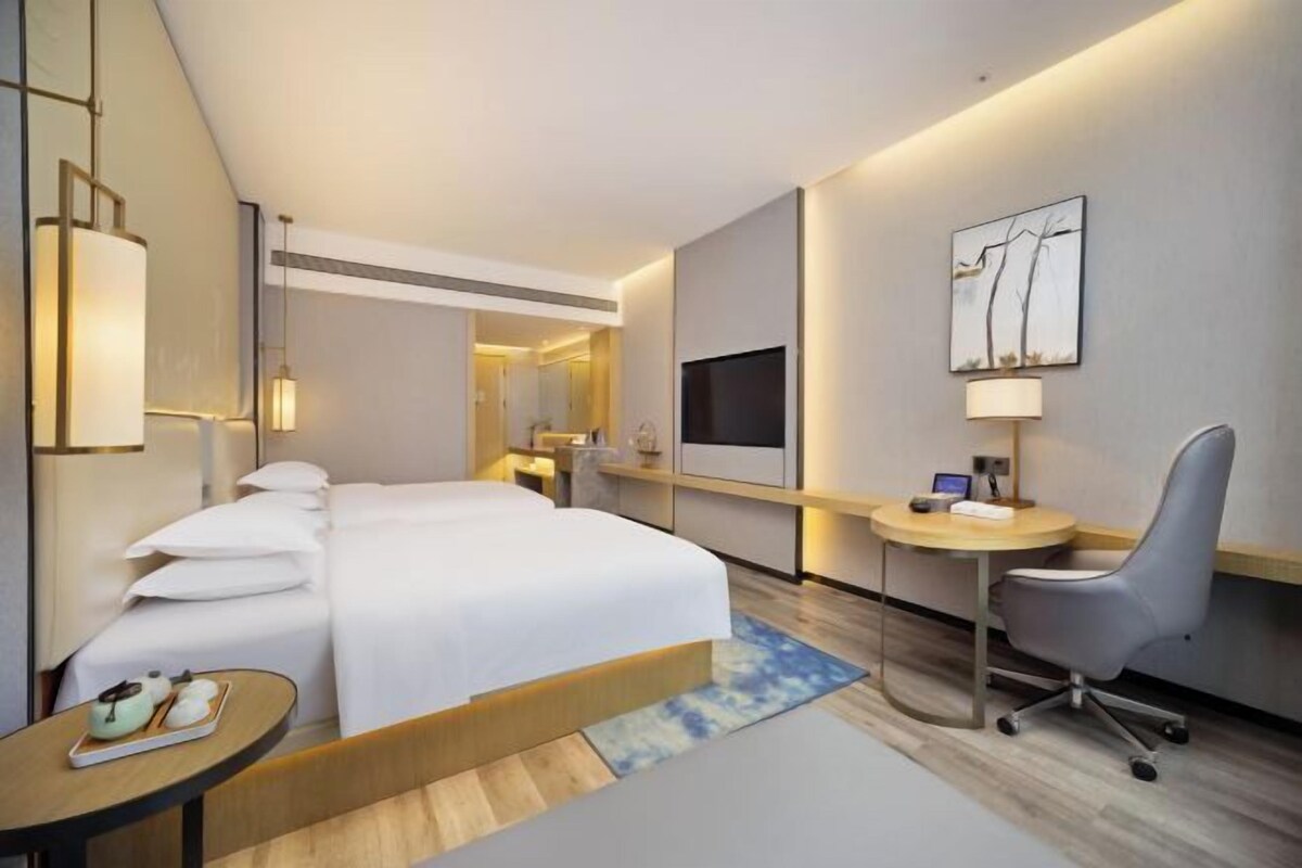 A hotel room with a bed, desk, chair, TV, and artwork.