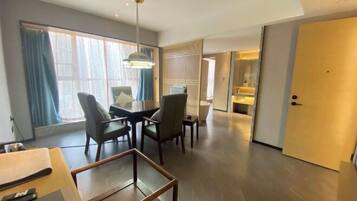 Una habitación de hotel moderna con comedor, un ventanal con cortinas, una puerta y un pasillo que conduce a un baño.