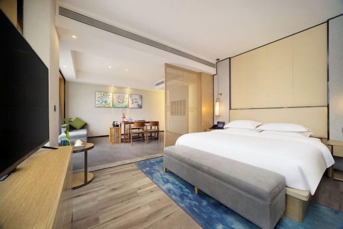 A hotel room with a large bed, a desk with a TV, a dining area with chairs, and artwork on the walls.