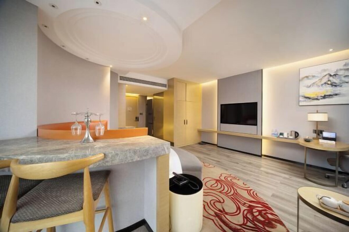 A modern hotel room with a marble dining table, wooden chairs, a TV, a desk with a computer, and a large painting on the wall.