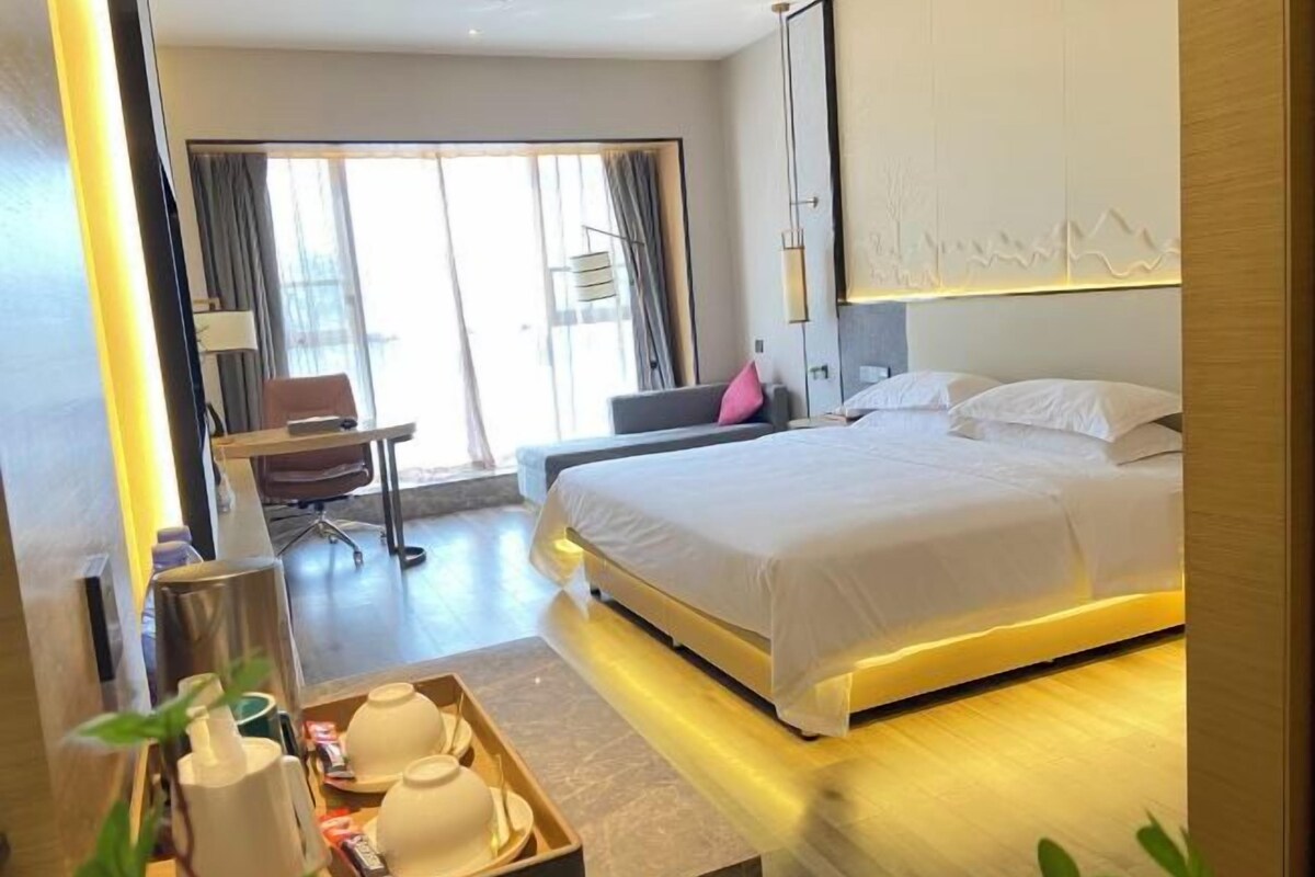 A hotel room with a bed, a desk, a chair, a window with curtains, and a wall with a light fixture.