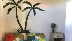 Een slaapkamer met een bed voorzien van kussens met een tropisch thema, een nachtkastje met een lamp en een houten dressoir.