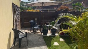 Een patio met een betonnen vloer, een houten omheining en een tafel met stoelen. Er staan potplanten en een palmboom op de achtergrond.