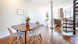 Un comedor con una mesa de madera y sillas blancas, una planta en maceta y una escalera que sube a un altillo.
