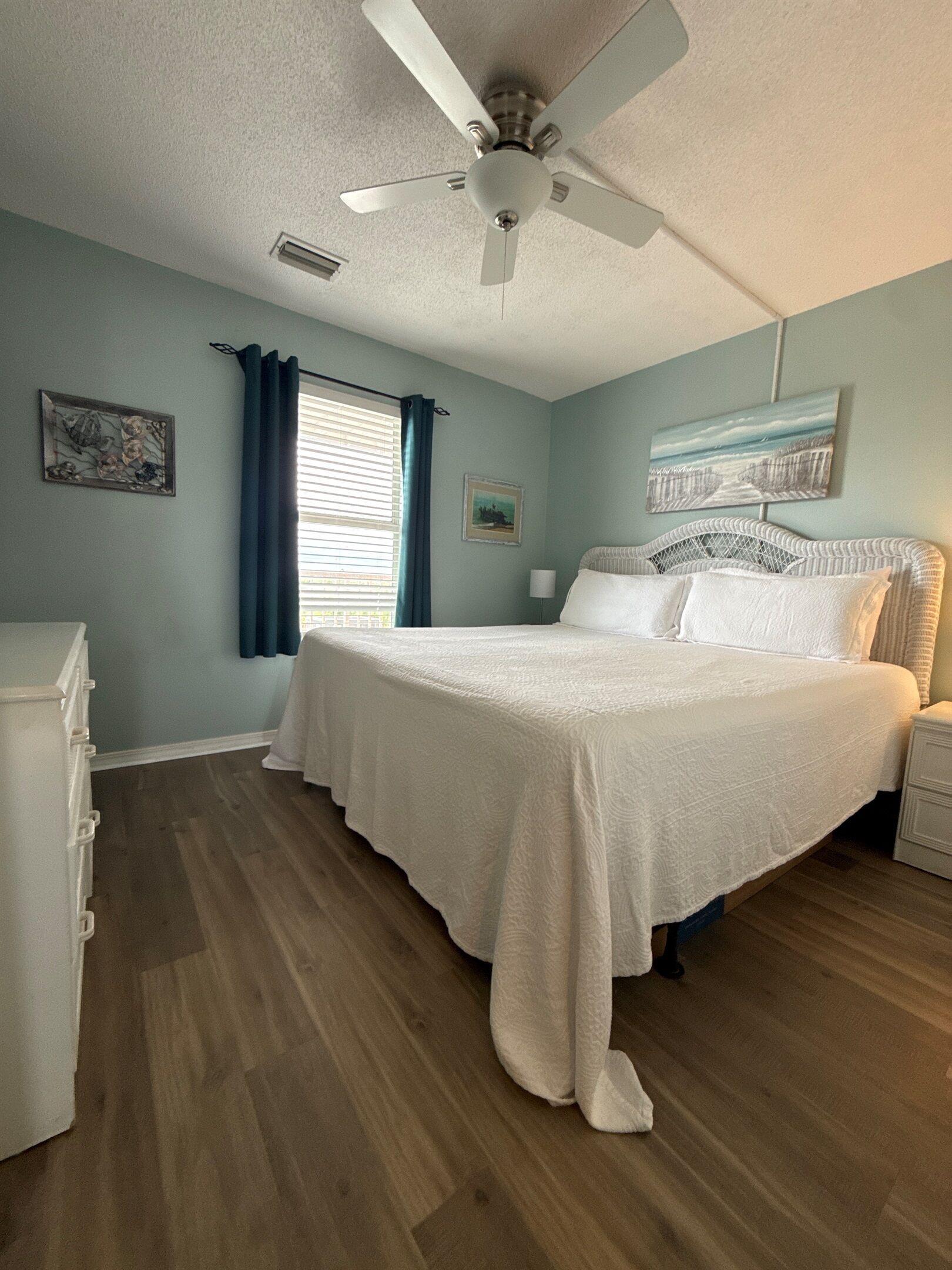 Un dormitorio con cama, ventilador de techo, ventana con cortinas y obras de arte en las paredes.