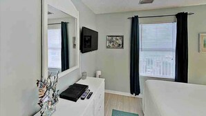 A bedroom with a large mirror, a bedside table with a remote control, a small decorative vase, and a window with curtains.
