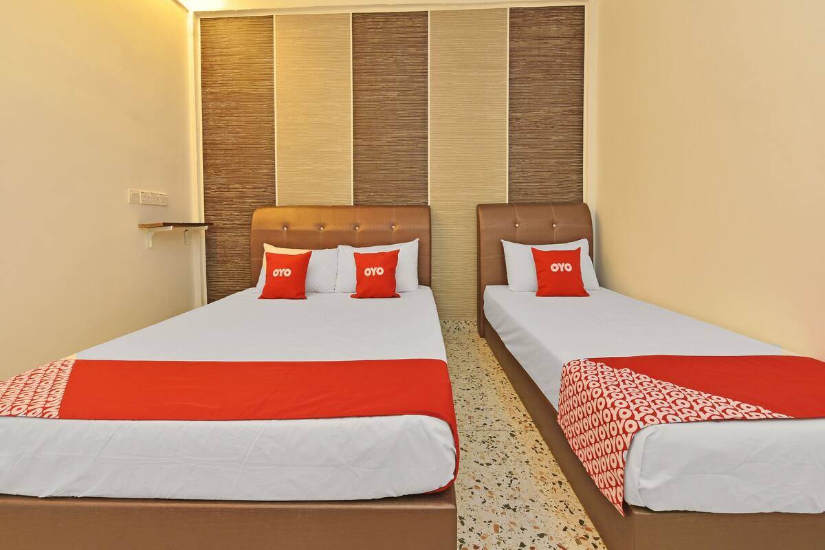 Une chambre d’hôtel avec deux lits, chacun recouvert d’une couvre-lit rouge et blanche assortie à des coussins du même style. Les têtes de lit sont recouvertes d’un tissu marron, et le mur derrière les lits est décoré de panneaux en bois.
