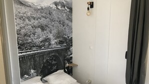 Un dormitorio con cama, ropa de cama blanca, una mesita de noche, una lámpara, un interruptor en la pared y una cortina.