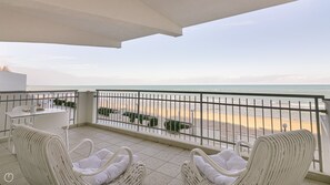 Een balkon met witte rieten stoelen, een tafeltje en uitzicht op het strand.