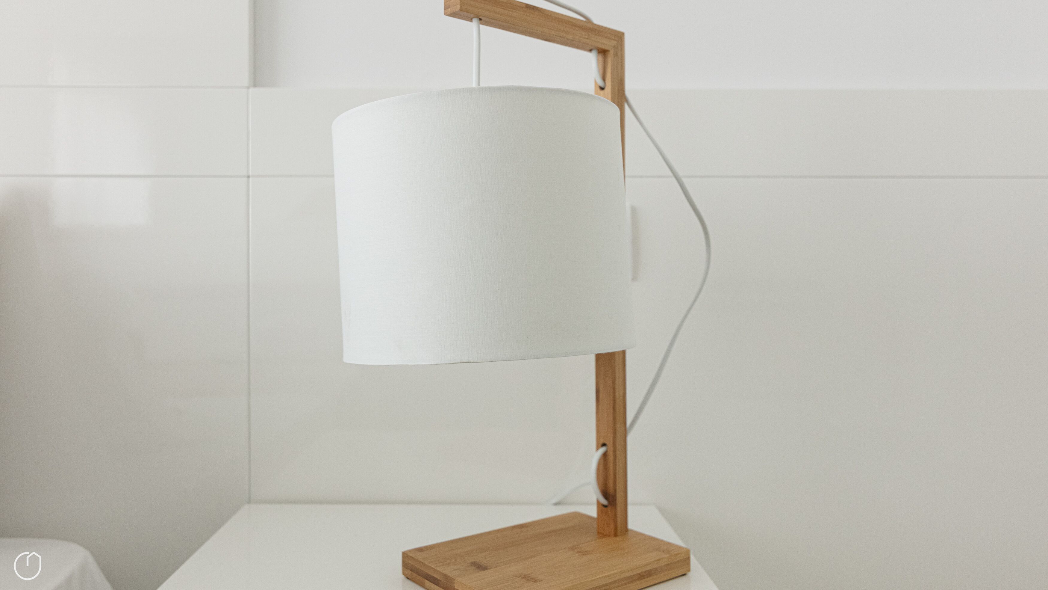 A modern desk lamp with a wooden base and a cylindrical shade, placed on a white surface against a white tiled wall.