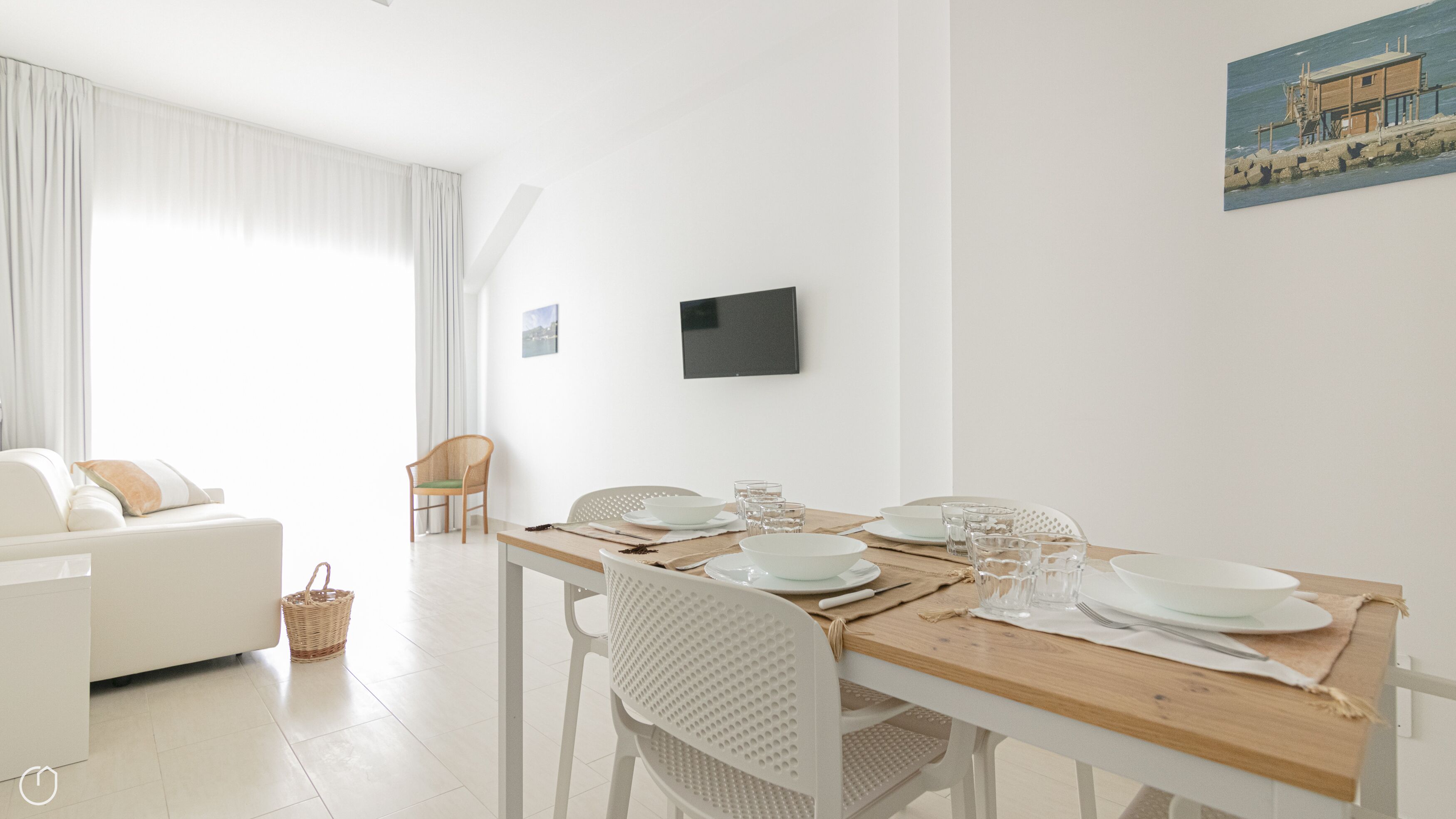 Una sala de estar moderna y luminosa, con un sofá blanco, una mesa de comedor dispuesta con platos y vasos, y un ventanal con cortinas de gasa.